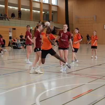 Joueuse du HBC Moudon en orange tentant un tir malgré les défenseuses numéros 6 et 3 du TV Birsfelden en bordeaux, avec des spectateurs sur la galerie