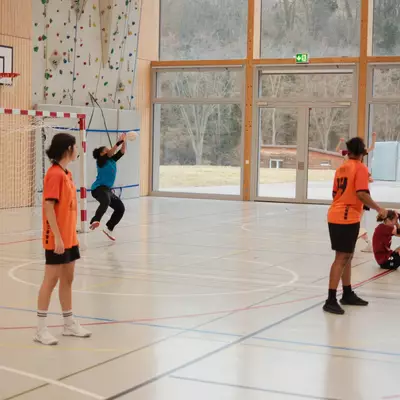 Gardienne du HBC Moudon en maillot bleu sautant pour capter le ballon devant le but, avec des coéquipières en orange observant la scène