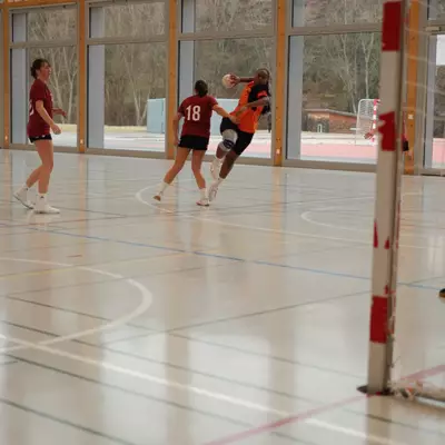 Joueuse du HBC Moudon en maillot orange tirant au but face à une défenseuse du TV Birsfelden numéro 18 en maillot bordeaux, avec la gardienne prête à intervenir