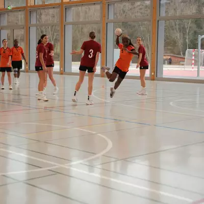 Joueuse du HBC Moudon en maillot orange s'envolant pour tirer au but, évitant les défenseuses du TV Birsfelden en maillot bordeaux