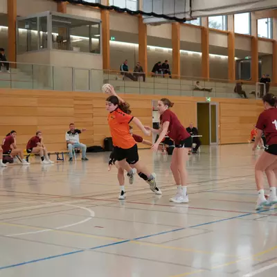 Joueuse du HBC Moudon en maillot orange sautant pour tirer, face aux défenseuses du TV Birsfelden en bordeaux, avec des spectateurs sur la galerie