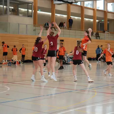 Joueuse du HBC Moudon en orange sautant pour tirer par-dessus les défenseuses numéros 10 et 3 du TV Birsfelden en bordeaux, avec le public sur la galerie
