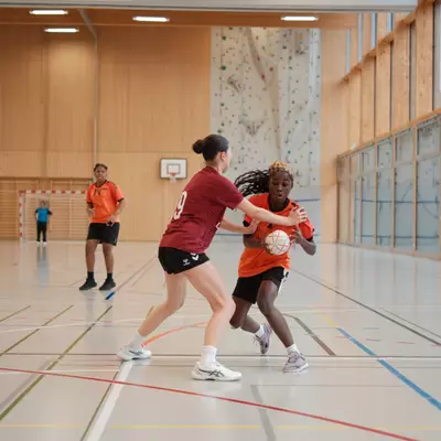 Joueuse du HBC Moudon en maillot orange protégeant le ballon face à une défenseuse numéro 9 du TV Birsfelden en bordeaux