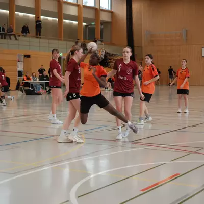 Joueuse du HBC Moudon en orange tentant un tir malgré les défenseuses numéros 6 et 3 du TV Birsfelden en bordeaux, avec des spectateurs sur la galerie