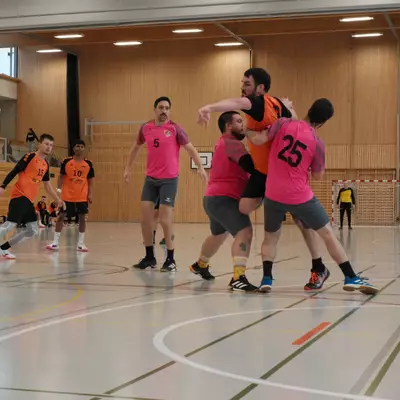 Joueur du HBC Moudon en orange retenu par deux défenseurs du RSB/Yverdon numéros 5 et 25 en rose, avec des coéquipiers observant la scène