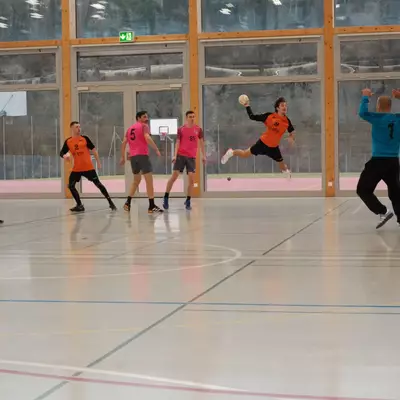 Joueur numéro 8 du HBC Moudon en orange s'envolant pour tirer au but, face au gardien du RSB/Yverdon numéro 1 en bleu, avec plusieurs joueurs des deux équipes observant l'action