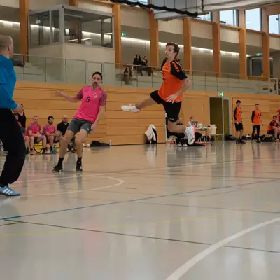 Joueur du HBC Moudon en orange sautant pour tirer, face à l'arbitre en bleu et un défenseur du RSB/Yverdon numéro 5 en rose, avec des spectateurs sur la galerie