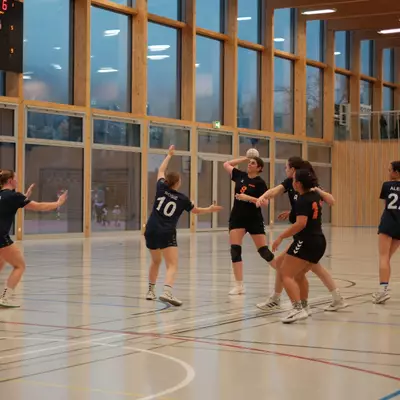 Joueuse numéro 8 du HBC Moudon en noir armant un tir entourée de défenseuses du NHB La Côte en bleu marine, avec le tableau d'affichage visible