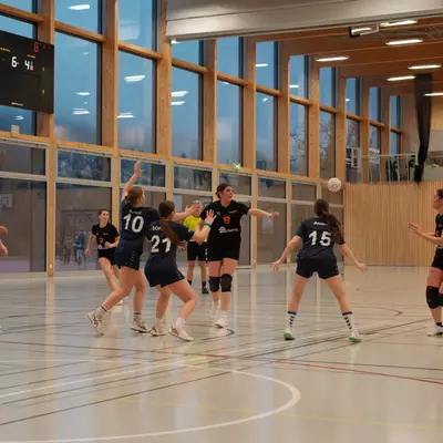 Joueuse numéro 8 du HBC Moudon en noir passant le ballon par-dessus la défense du NHB La Côte, avec le score 6-4 au tableau d'affichage