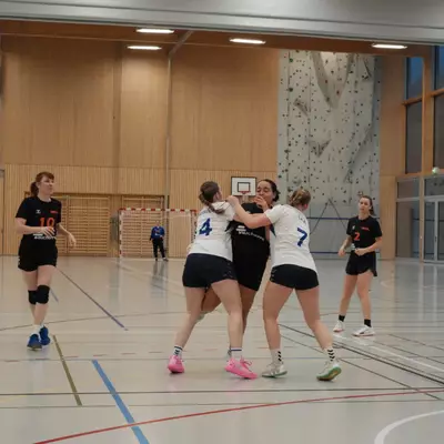 Deux joueuses du NHB La Côte numéros 4 et 7 en blanc bloquant une joueuse du HBC Moudon en noir, avec d'autres joueuses observant l'action