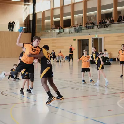 Orange player number 13 leaps to shoot during a youth boys handball match while a black-and-yellow defender tries to block, with other players visible and spectators watching from an upper gallery