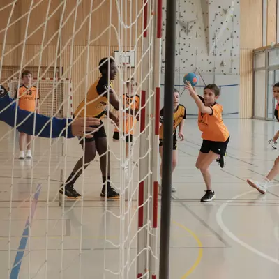View through the goal net of an orange youth handball player raising the ball to shoot, with black-and-yellow opponents nearby, the blue goalkeeper on the left, and a climbing wall with large windows in the background
