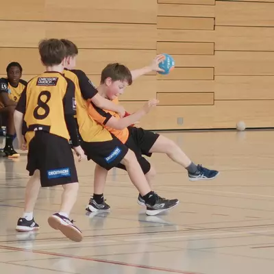 An orange youth handball player shoots while under pressure from black-and-yellow defender number 8, with seated players and a spectator watching from the sideline