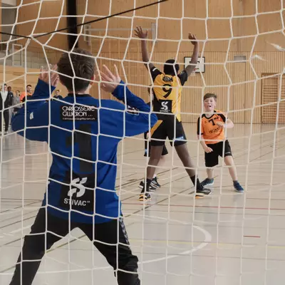 View through the goal net of the blue goalkeeper raising both arms to save as black-and-yellow player number 9 leaps with arms raised and orange player number 1 looks on during a youth handball match