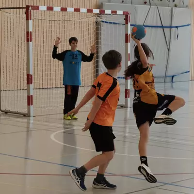 Blue goalkeeper number 12 raises both arms in the goal as a black-and-yellow youth player leaps to shoot during a handball match, with an orange player watching and a climbing wall visible through large windows in the background