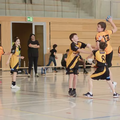 Orange player number 13 leaps to shoot past two black-and-yellow defenders during a youth handball match, with orange teammates number 15 and 7 visible on the left and a coach watching in the background