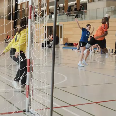 An orange player jumps to shoot past a blue defender near the goal while the goalkeeper in yellow moves to block, seen through the goal post frame with spectators watching from a gallery above