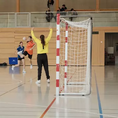 An orange handball player shoots toward goal while the goalkeeper in yellow raises both arms to block, viewed from beside the goal frame with a blue defender nearby