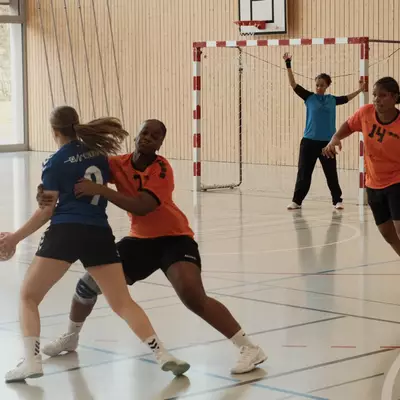 An orange player closely marks a blue ball-carrier near the handball goal, with the goalkeeper in blue watching from behind and orange number 14 providing defensive support on the right