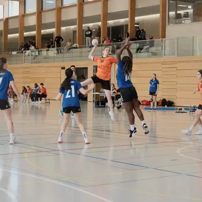 An orange player leaps high with the ball above two blue defenders during a handball match, with spectators and players from both teams watching in a sports hall with an upper gallery