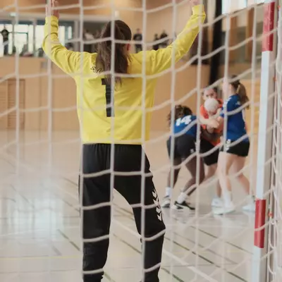 View through the goal net of a goalkeeper in yellow raising her arms while a blue player number 14 holds the ball close to the goal post during a handball match