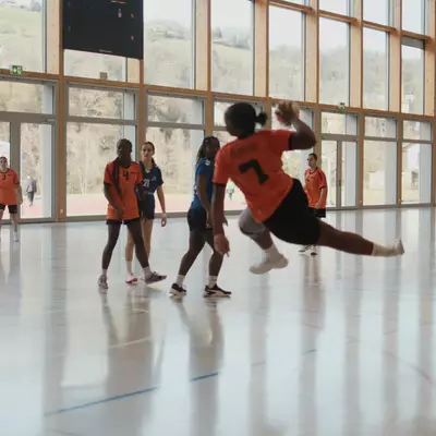 Orange player number 7 leaps to throw the ball during a women's handball match in a modern indoor sports hall with large windows overlooking a hillside