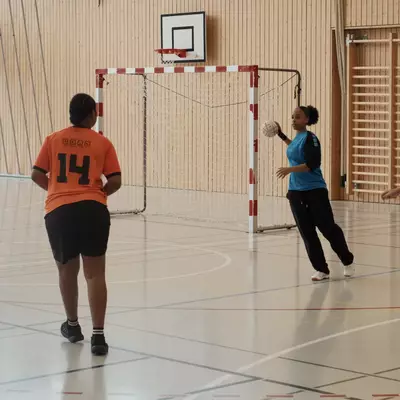 Orange player number 14 walks toward the handball goal while a blue-jerseyed player holding the ball prepares to pass to a running teammate during a training exercise