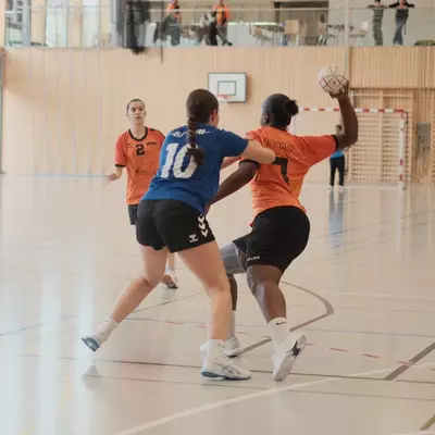 An orange handball player shields the ball overhead while a blue defender number 10 contests possession, with another orange player number 2 watching from behind in an indoor sports hall
