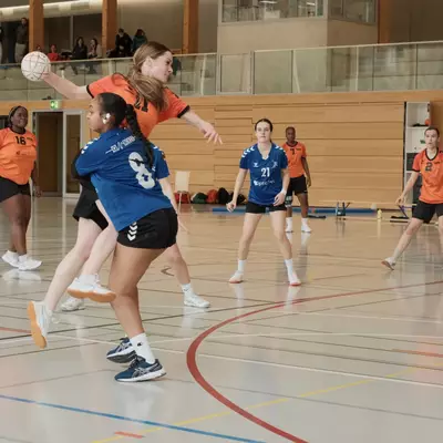 An orange player leaps and extends her arm to pass the ball over blue defender number 8 during a women's handball match, with spectators watching from an upper gallery