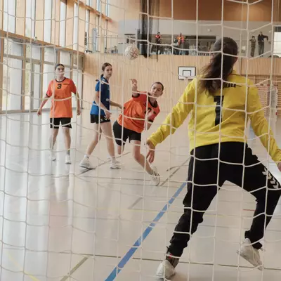 View from behind the goal net of a goalkeeper in yellow in a defensive stance as an orange player shoots the ball, with several players from both teams visible on the court