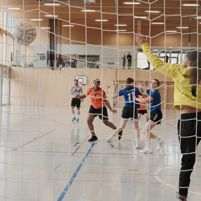 View from behind the goal net of a handball sailing into the top corner as the goalkeeper in yellow stretches to block it, with blue player number 1 having just shot and multiple players positioned on the court