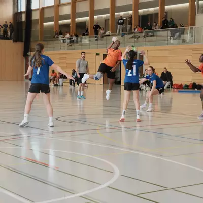 An orange player jumps high with the ball above outstretched arms of blue defenders during a handball match, with players from both teams positioned around the play and spectators watching from an upper gallery