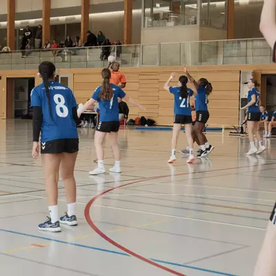 An orange player rises above blue defenders with the ball ready to shoot as multiple opponents raise their arms to block, with a crowd of spectators watching from an upper gallery