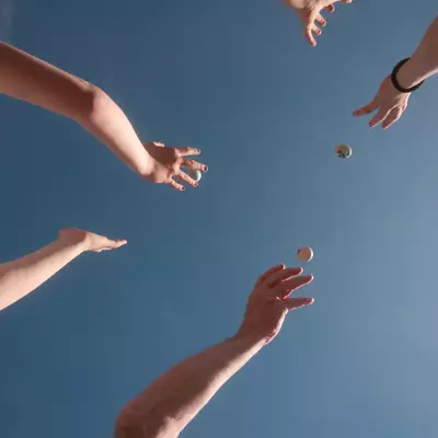 Low-angle view of three people's arms and hands reaching up toward the sky, throwing and catching Easter eggs. A child in a white t-shirt is visible on the left. Two eggs are mid-air at the center of the image. The background is a clear, uniform blue sky.