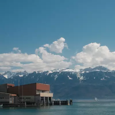 The CGN harbor building at Ouchy pier extending over Lake Geneva, with a small white boat docked alongside, a sailboat in the distance, and snow-covered Alps under a blue sky.