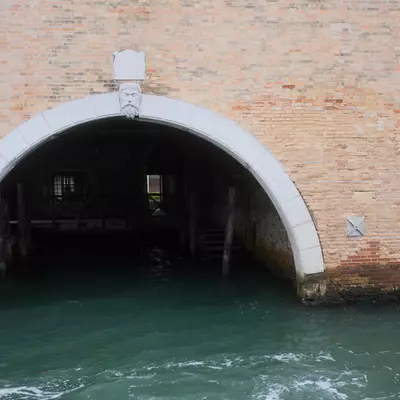 Arched water gate of a brick building opening directly onto a turquoise canal in Venice, a carved stone face above the arch keystone