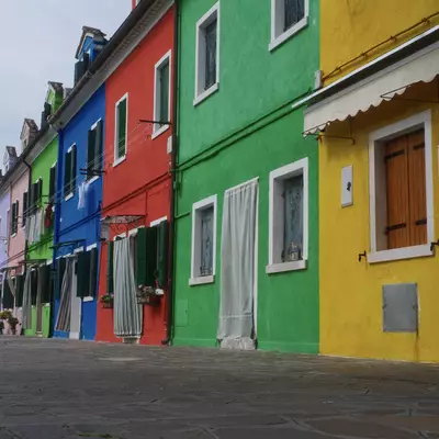 Row of brightly coloured Burano houses in blue, red, green and yellow along a wide cobblestone street