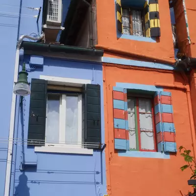 Two adjacent house facades in Burano painted periwinkle blue and burnt orange, with multicoloured striped shutters and a casabepi.it sign