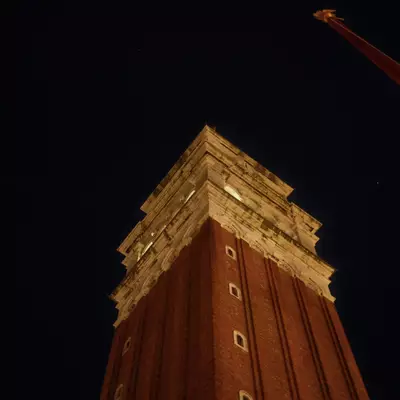 Top of the Campanile di San Marco illuminated at night against a black sky, a red construction crane visible in the upper right corner