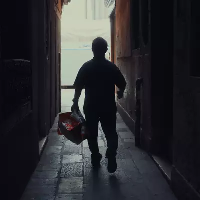 Silhouette of a person carrying a bag walking down a narrow dark Venetian calle toward bright light at the far end
