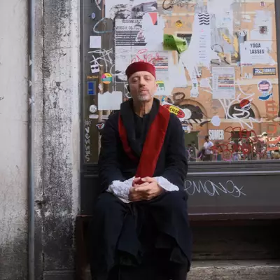 A man in traditional Venetian theatrical costume — black robes, red beret and red velvet stole — sitting on a step in front of a wall covered with stickers and posters