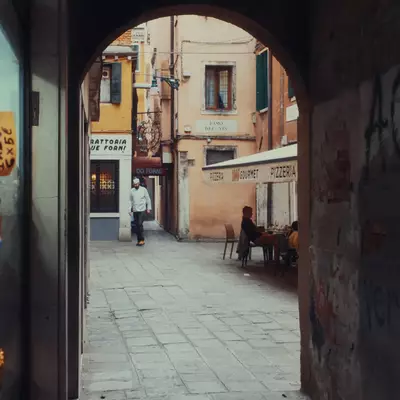 View through a stone archway into a Venetian campo, a chef in white uniform walking past, people sitting at outdoor pizzeria tables, graffiti on the arch walls