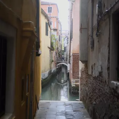 Narrow Venetian calle opening onto a small canal with a stone footbridge, a solitary figure crossing in the distance