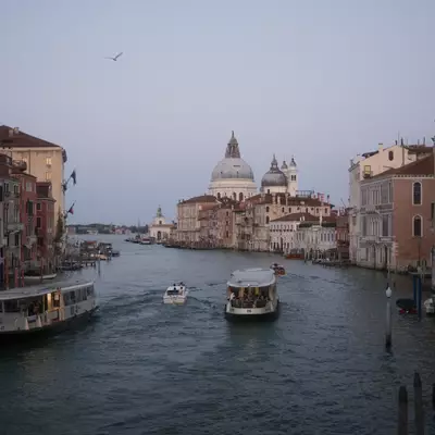 View of the Grand Canal in Venice at dusk, with vaporetti and a small motorboat navigating the waterway. Historic Venetian palaces line both sides of the canal, and the domed basilica of Santa Maria della Salute rises in the background under a hazy blue-grey sky. A seagull flies overhead.
