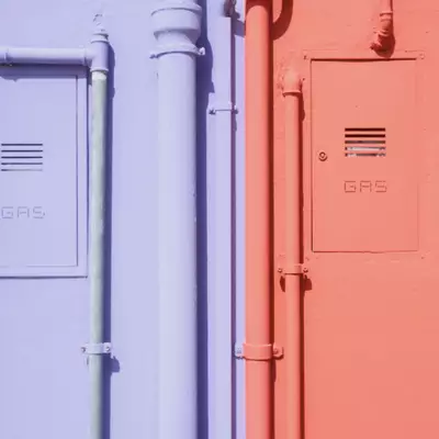 Close-up of two adjacent Burano house walls painted lavender and coral, each with a gas meter box and pipes painted in the matching wall colour