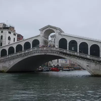 The Rialto Bridge spanning the Grand Canal in Venice, viewed from water level, with boats moored below under an overcast sky