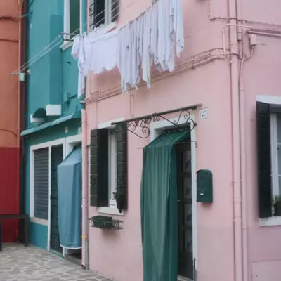 Pink and teal painted houses on Burano island, white laundry hanging on a line between them, green curtains in the doorways