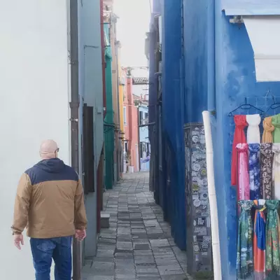 A man walking away into a colourful narrow alley in Burano, scarves for sale hanging on the right