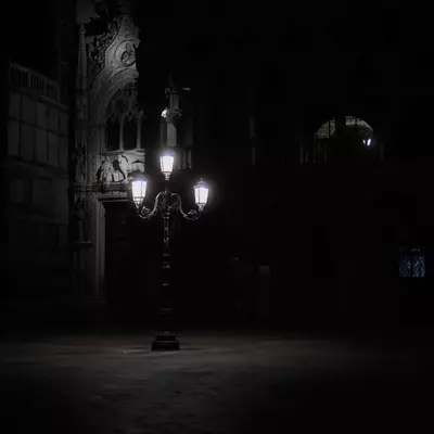 A wrought iron lamp post with three glowing lanterns illuminating the facade of the Doge's Palace at night, a winged lion sculpture visible in the darkness behind