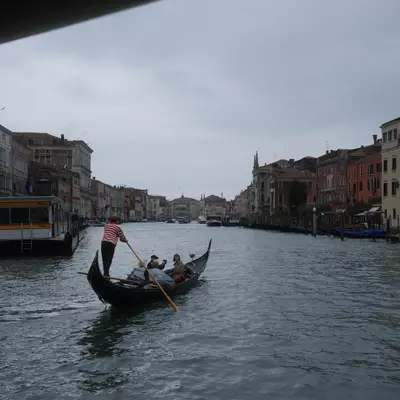 Gondolier in a striped shirt rowing tourists along the Grand Canal in Venice, colourful palace facades lining both banks under an overcast sky